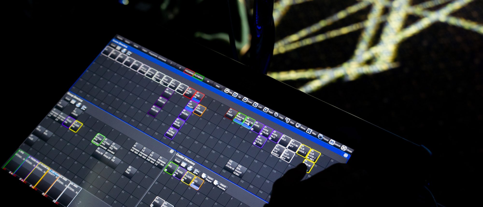 A hand on a lighting console touchscreen with coloured cue buttons, light beams crossing in the background