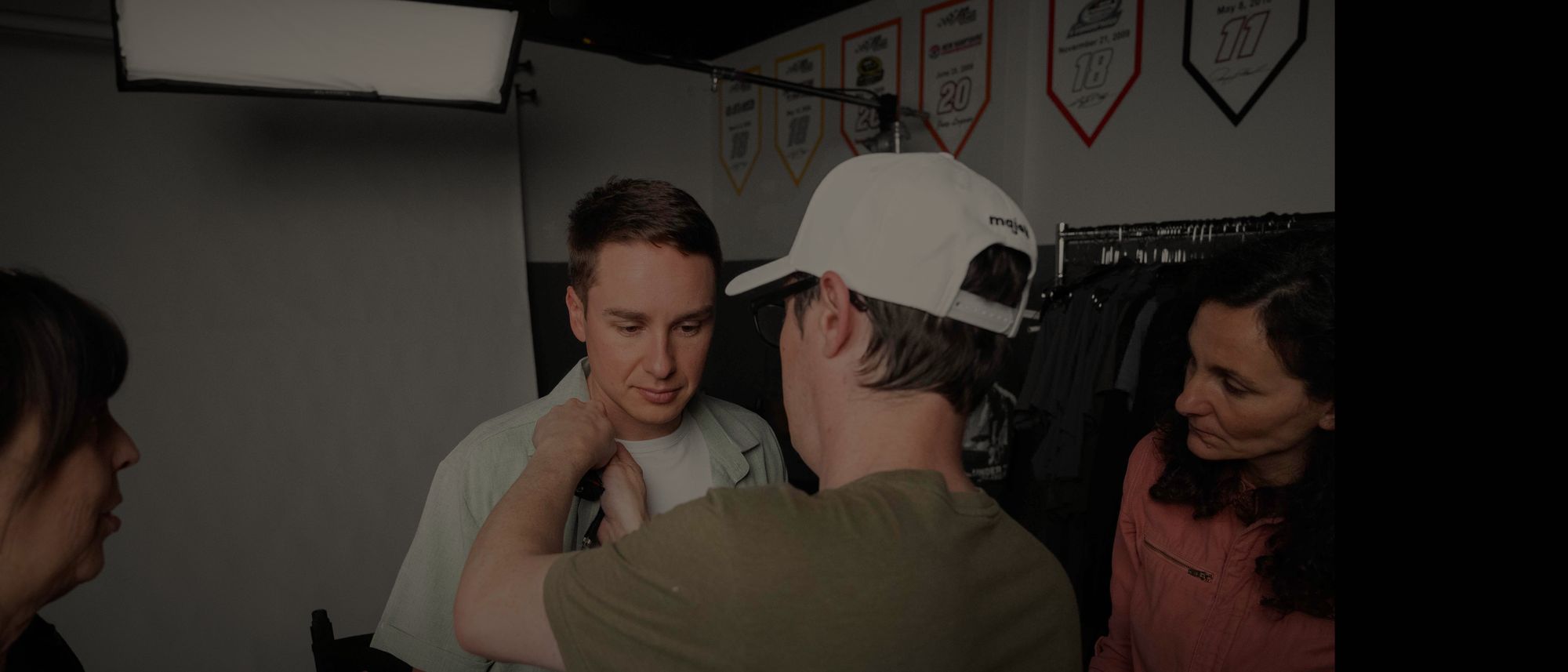 Christopher Bell in a green shirt has his collar adjusted by a crew member while two others look on, a lit studio behind them
