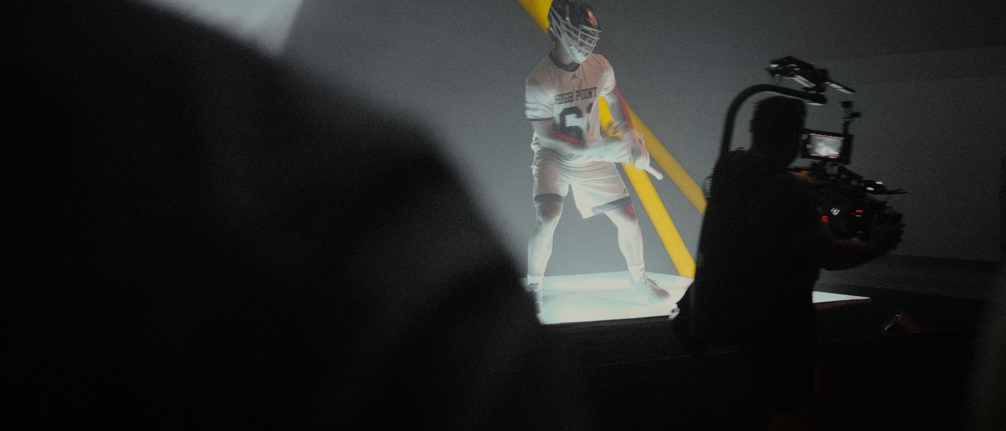 A lacrosse player in a beam of light on a dark set while a camera operator films him on a gimbal