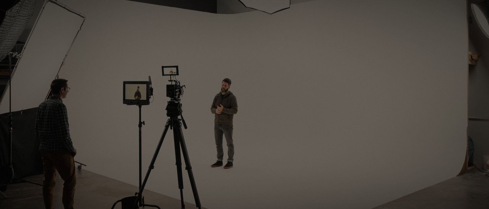 A camera on a tripod facing a presenter on a large white cyc, with a crew member watching from the side