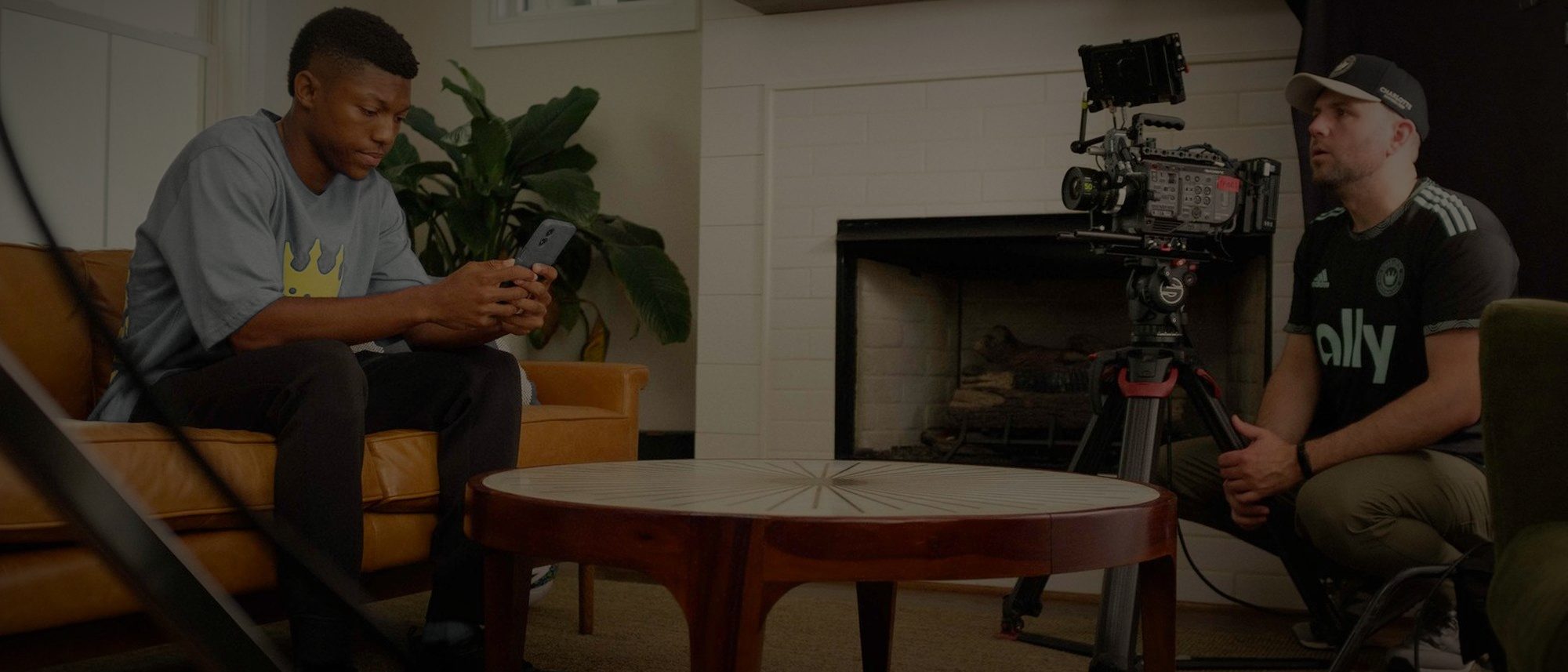 Two people seated in a living room set during an interview setup, with a camera on sticks between them.