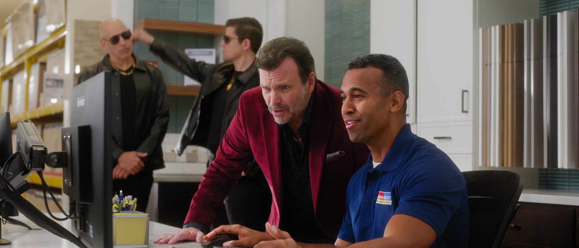 A store associate at a computer shows a price to a man in a red velvet jacket while two men in sunglasses wait behind